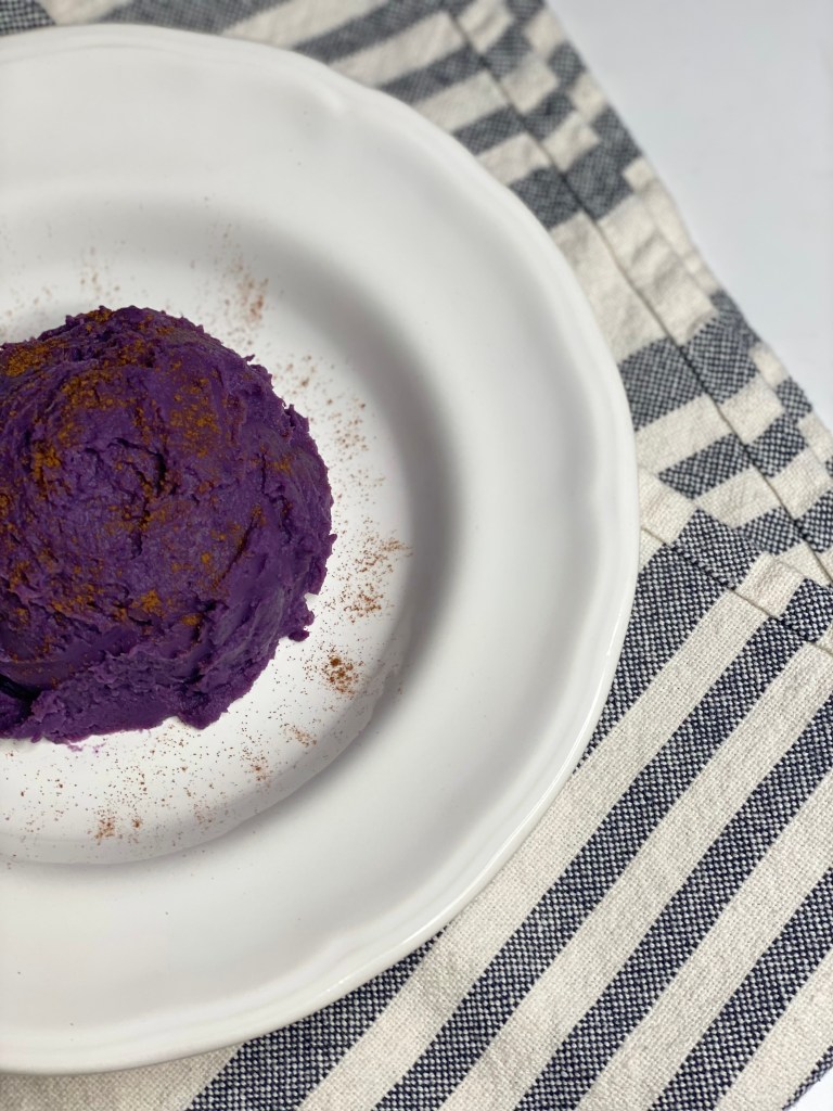 Large scoop of sweet purple potato mash in a bowl with a striped dish towel.