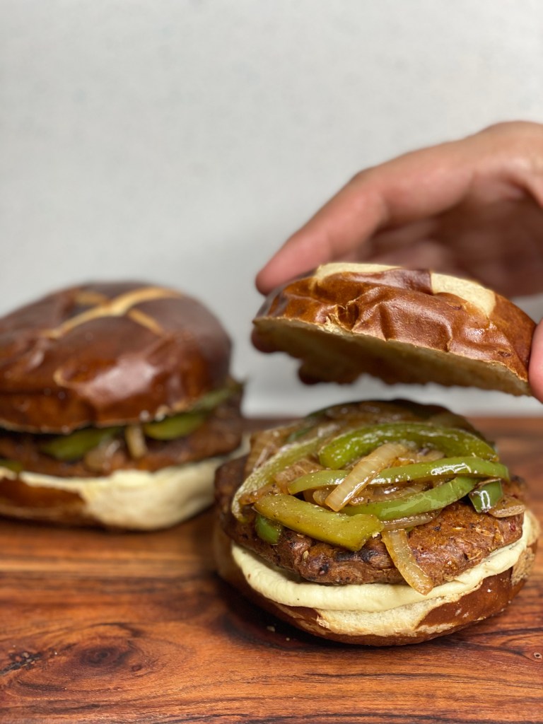 A hand assembling a vegan spicy sausage burger bun with peppers, onions and cheese sauce on a pretzel bun.