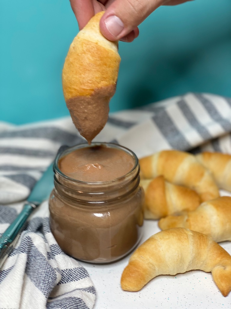 Hand dipping a pastry into a jar of vegan chocolate nutella spread.