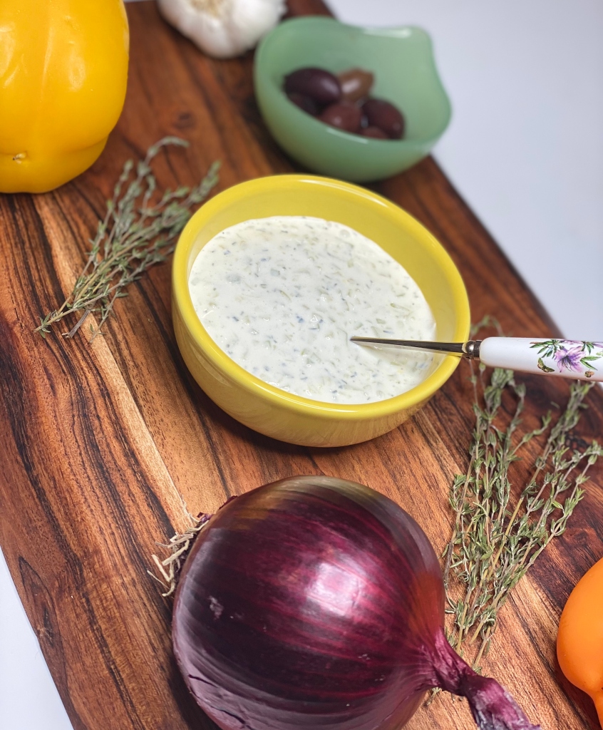 Creamy tzatziki sauce in a yellow bowl with thyme, olives, garlic, bell peppers and red onion.